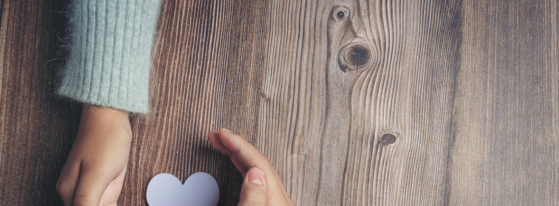 Hände halten ein Herz aus Papier auf einem Untergrund aus Holz