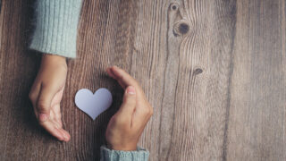 Hände halten ein Herz aus Papier auf einem Untergrund aus Holz