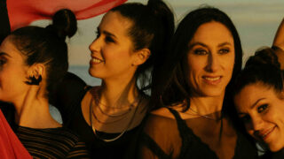 Vier Künstlerinnen, die beim Women in Emotion Festival auftreten, stehen am Meer und lassen ein rotes Tuch über den Köpfen schweben