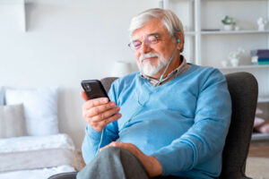 Ein Senior sitzt mit dem Smartphone in der Hand im Sessel und hat Kopfhörer im Ohr