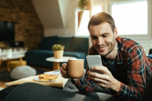 Ein Mann liegt auf dem Bauch auf der Couch und hält in der einen Hand eine Tasse, in der anderen ein Smartphone