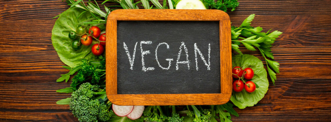 Eine kleine Tafel mit der auf Kreide das Wort vegan geschrieben wurde und im Hintergrund liegen Gemüse und Kräuter