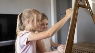 Zwei Kinder stehen an einer Staffelei und malen auf einem darauf gespannten Papier
