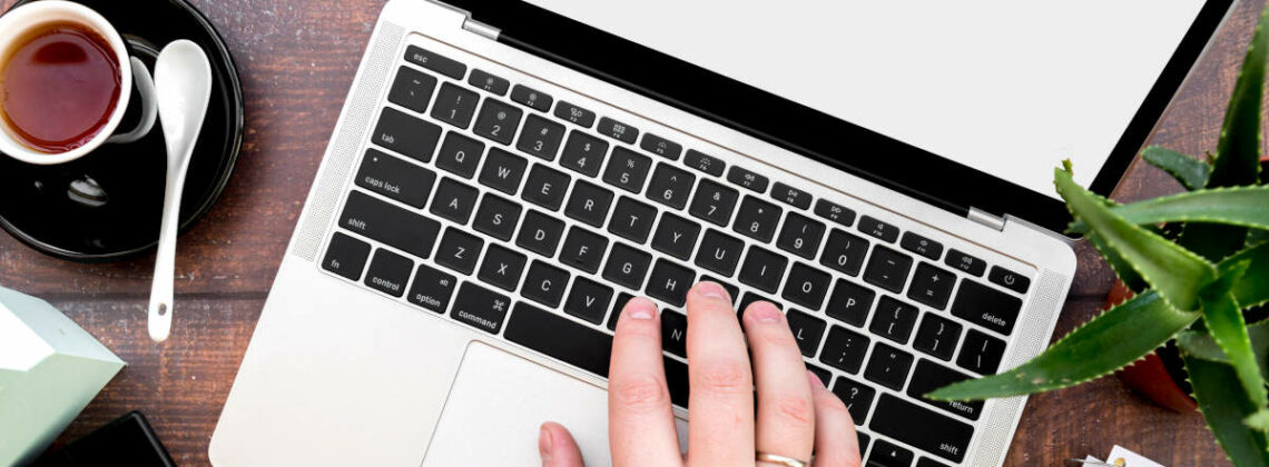 Eine Hand liegt auf einem Laptop, daneben liegt ein Taschenrechner sowie ein Notizblock