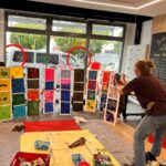 Eine Künstlerin und Kinder bestücken einen Aufsteller im Communitybereich der Sparkasse Bremen mit selbst gemalten Bildern