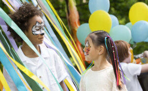 Ein Junge und ein Mädchen sind bunt geschminkt. Sie stehen inmitten bunter Bänder. Im Hintergrund sind Luftballons. Symbolbild für den Vegesacker Kindertag