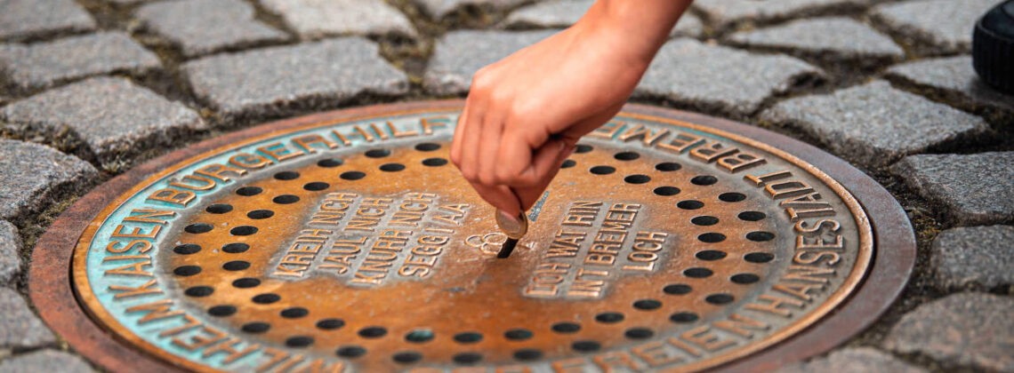 Nahaufnahme der Spendenbox "Das Bremer Loch" mit einer Hand, die Geld in den Schlitz wirft