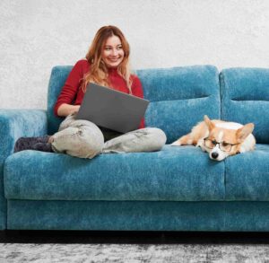 Das Foto zeigt eine lächelnde Frau auf einem Sofa mit einem Laptop, daneben liegt ein Hund, der eine Brille trägt.