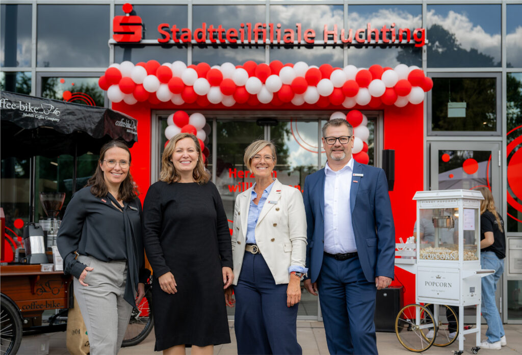 Gruppenfoto mit der Generalbevollmächtigten Sonja Kastin mit Franziska Heider, Sabine Hinne und Mathias Engelken (v.l.) von der Stadtteilfiliale Huchting bei der Eröffnung, im Hintergrund die Filiale mit Ballons und Popcornmaschine.