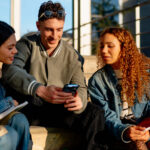Drei junge Menschen sitzen auf einer Treppe und gucken gemeinsam auf ein Smartphone. Symbolbild für die neue Bremer App Museenquiz