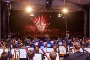 Blick von der Bühne auf das Orchester bei Nacht, im Hintergrund ein farbenfrohes Feuerwerk über dem See bei „Musik und Licht am Hollersee“