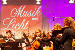 Jugendorchester spielt auf einer beleuchteten Bühne bei „Musik und Licht am Hollersee“, im Hintergrund ist der Schriftzug der Veranstaltung projiziert