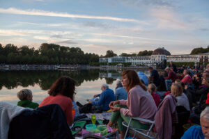 Menschen sitzen am Ufer des Hollersees mit Decken und Klappstühlen, warten auf den Konzertbeginn bei „Musik und Licht am Hollersee“