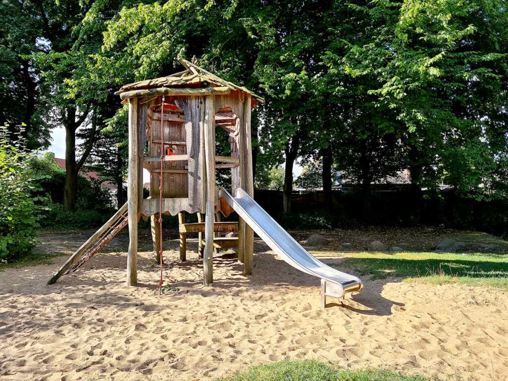 Hölzernes Spielhaus mit rustikalem Dach auf Stelzen, ausgestattet mit einer Edelstahlrutsche, einem roten Kletterseil und einer Kletterrampe. Das Spielgerät steht auf einem Sandboden, im Hintergrund dichter Baumbestand, der Schatten spendet.