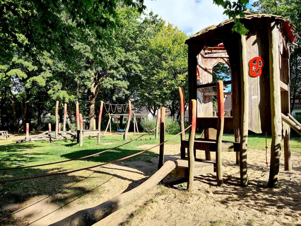 Blick vom Spielhaus auf einen großen Naturspielplatz mit vielen Holzspielgeräten. Im Vordergrund ein Balancierbereich mit Seilen und Baumstämmen, im Hintergrund eine Vogelnestschaukel und weitere Kletterelemente. Umgeben von großen, schattenspendenden Bäumen.