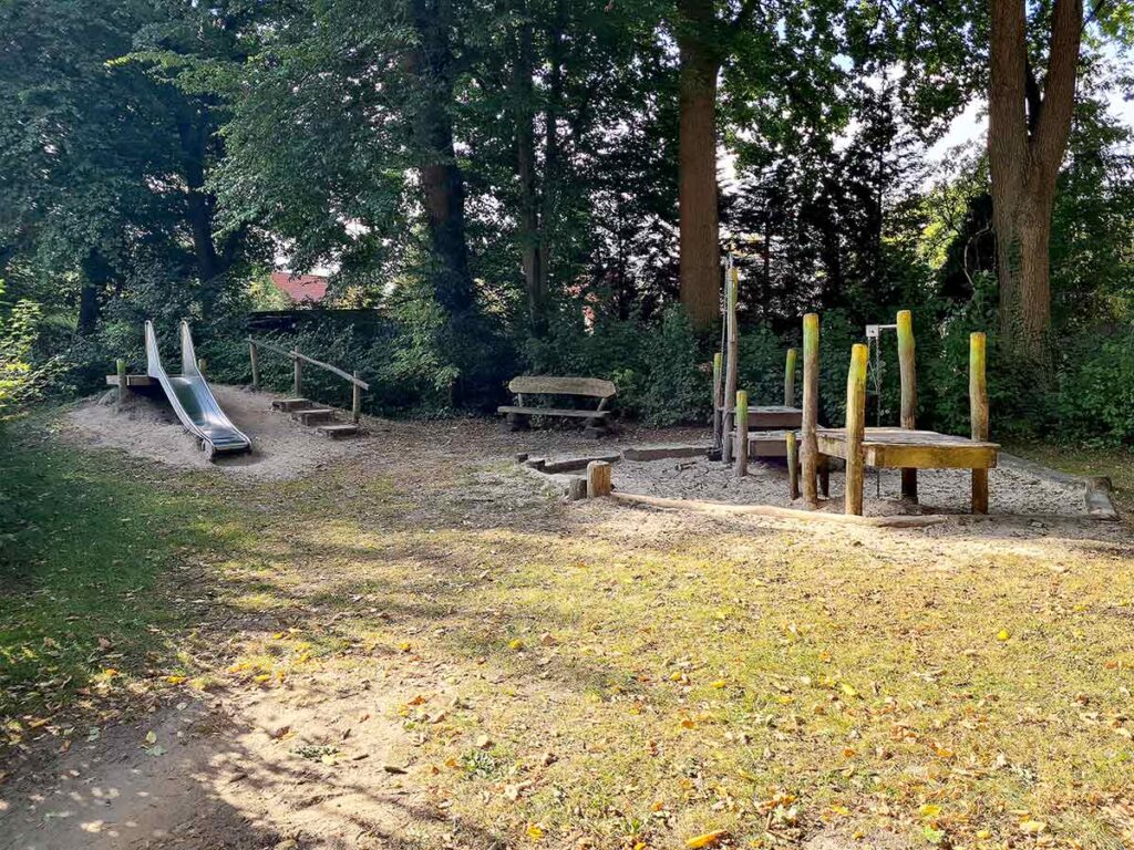 Übersicht über einen ruhig gelegenen Teil des Apoldi-Spielplatzes mit einer kleinen Rutsche auf einem Hügel links und einem Wasserspielbereich mit Holzplattformen und Pumpe rechts. Dazwischen stehen Bänke unter hohen schattenspendenden Bäumen. Der Boden ist mit Sand und Gras bedeckt.