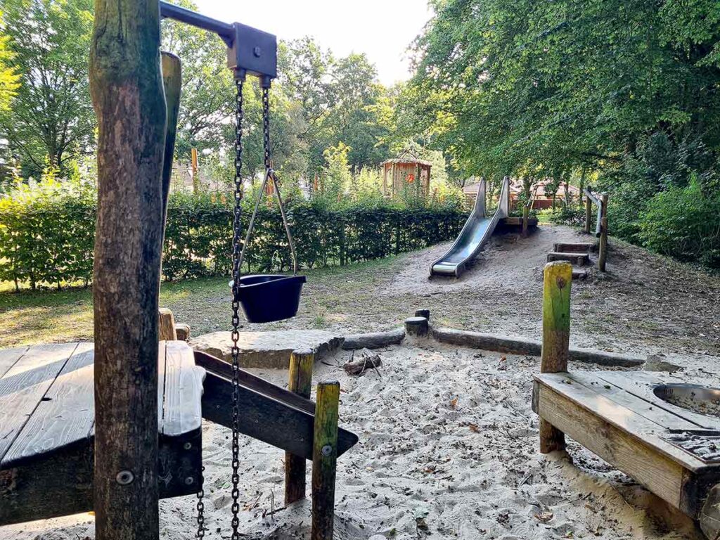 Blick auf den Wasser-Matschbereich des Apoldi-Spielplatzes mit einer schwarzen Schöpfkelle an einer Kette im Vordergrund. Dahinter eine kleine Edelstahlrutsche auf einem Hügel. Umgeben von Bäumen und Büschen bietet der Platz viel Schatten und Natur.