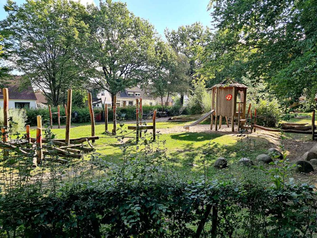 Übersicht über den gesamten Apoldi-Spielplatz in Vegesack. Zu sehen sind der zentrale Spielturm mit Rutsche sowie zahlreiche Kletterelemente aus Holz. Der Spielplatz liegt inmitten eines Wohngebiets und ist von Bäumen und Büschen umgeben, was viel Schatten und eine naturnahe Atmosphäre bietet.