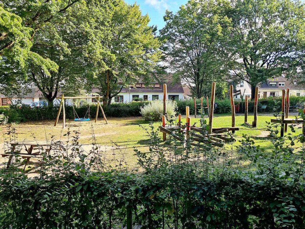 Blick auf die Kletterlandschaft und Nestschaukel des Apoldi-Spielplatzes in Bremen-Vegesack. Die Spielgeräte bestehen überwiegend aus naturbelassenem Holz und stehen auf einer großen, grünen Rasenfläche. Im Vordergrund ist eine Sitzbank zu erkennen, im Hintergrund befinden sich Wohnhäuser.