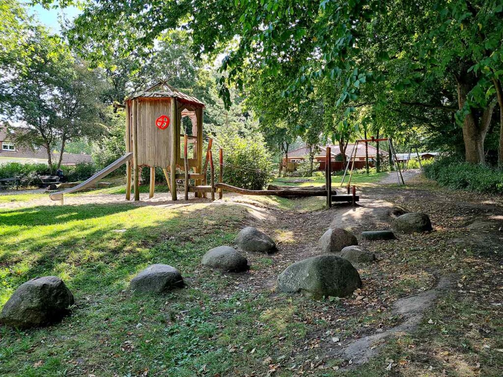 Naturspielplatz in Bremen-Vegesack mit Holzspielhaus samt Rutsche und Kletterzugang. Im Vordergrund liegen große Findlinge, die als Balanciersteine genutzt werden können. Im Hintergrund sieht man eine Doppelschaukel und viel Grün unter schattigen Bäumen.