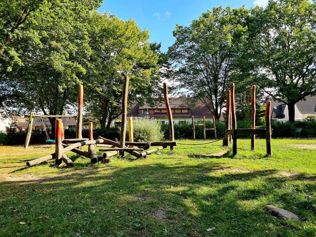 Große Balancierlandschaft aus naturbelassenem Holz auf einer grünen Wiese. Die Anlage besteht aus Baumstämmen, Seilen und Holzstegen zum Klettern und Balancieren. Im Hintergrund ist eine Vogelnestschaukel zu sehen, umrahmt von Bäumen und Wohnhäusern.