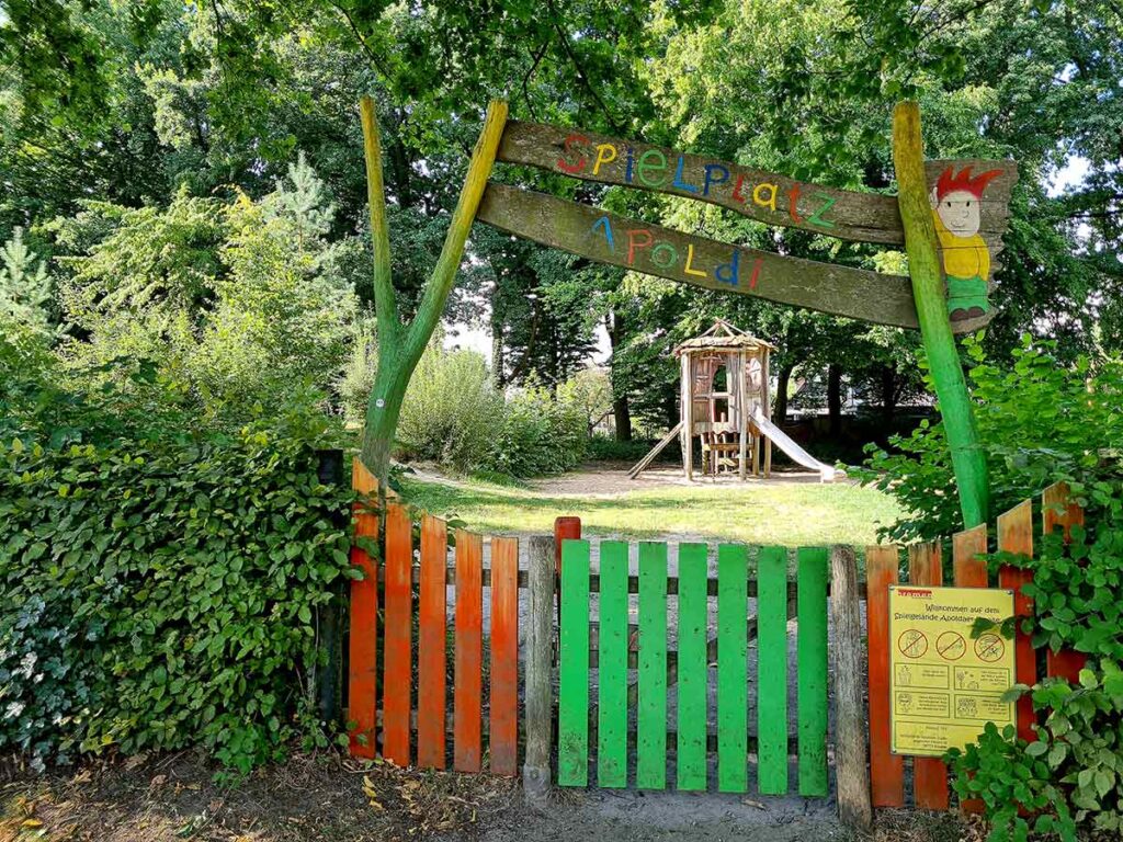 Bunter Eingangsbereich des Spielplatzes „Apoldi“ mit bemaltem Holztor in Rot-, Orange- und Grüntönen. Ein hölzernes Schild mit dem Namen „Spielplatz Apoldi“ und einer gemalten Figur ziert den oberen Bereich. Im Hintergrund ist ein hölzernes Spielhaus mit Rutsche zu sehen, umgeben von grünem Rasen und Bäumen.