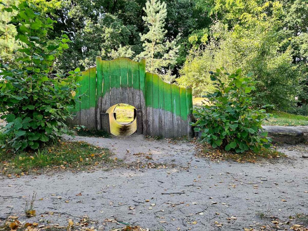 Grünes Spieltunnel-Element aus Holz auf dem Spielplatz Apoldi. In der Mitte ein runder Durchgang zum Durchkriechen, eingerahmt von bemalten Holzplanken in Grün- und Naturtönen. Umgeben von kleinen Büschen und Bäumen auf einem sandigen Untergrund.