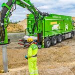 Ein grün lackiertes Baufahrzeug vonm Nehlsen hat einen großen Erdbohrer ausgefahren, der sich in den Sand bohrt. Ein Mitarbeiter in Arbeitskleidung beobachtet das.