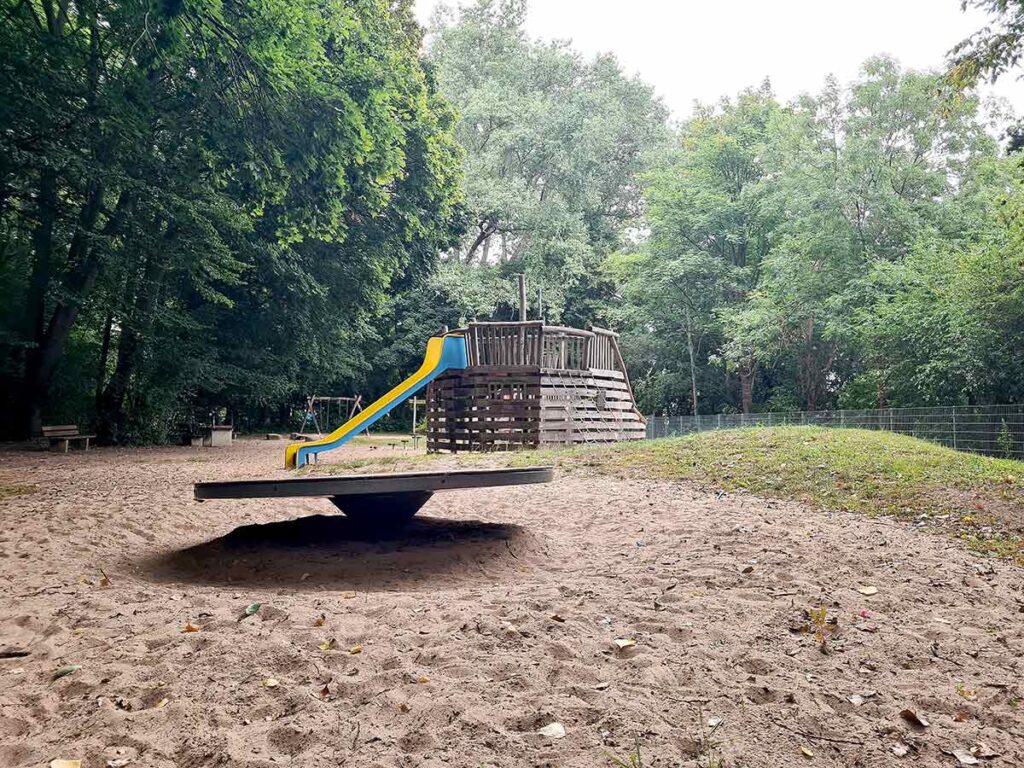 Übersicht über den bewaldeten Spielplatz mit Drehkreisel im Vordergrund und großem Holzspielschiff mit Rutsche im Hintergrund, eingebettet in eine naturnahe Sandlandschaft.