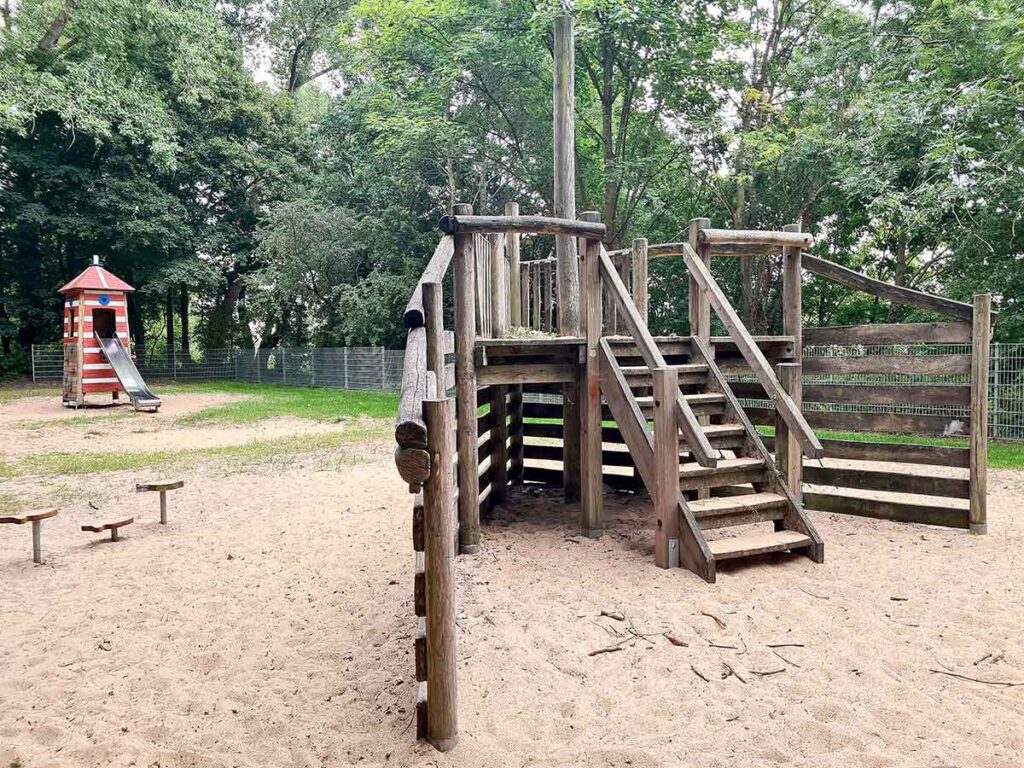 Nahaufnahme des hölzernen Piratenschiffs mit breiter Treppe; im Hintergrund ein rot-weiß gestreifter Leuchtturm mit Rutsche und weitere Spielgeräte auf sandigem Boden zwischen Bäumen.