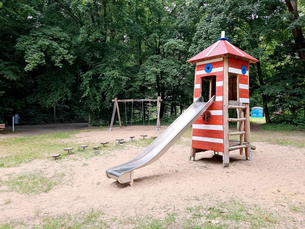 Roter Leuchtturm mit weißem Streifenmuster und Edelstahlrutsche auf dem Spielplatz im Dillener Park, daneben eine Holzschaukel und Balancierstangen, umgeben von Bäumen.