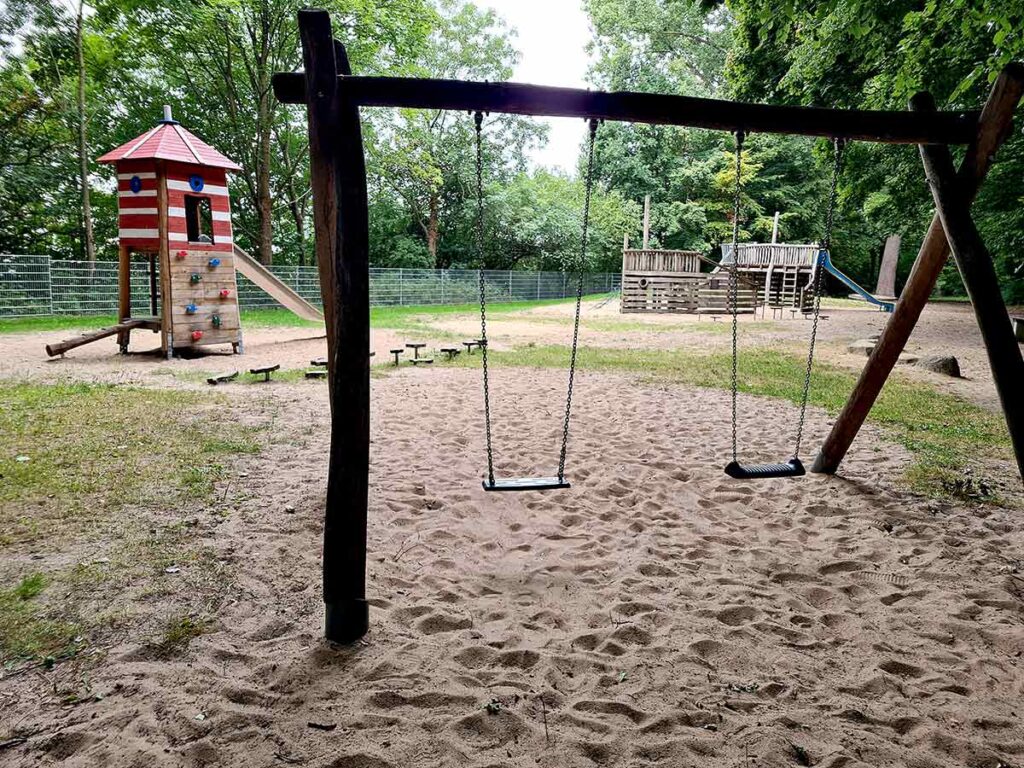 Blick über den Spielplatz im Dillener Park mit einer Holzschaukel im Vordergrund, einem roten Leuchtturm mit Rutsche und Kletterwand links sowie einem hölzernen Spielschiff im Hintergrund – alles umgeben von dichtem Baumbestand.