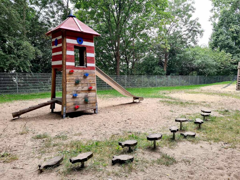 Roter Spiel-Leuchtturm mit weißem Streifenmuster, Edelstahlrutsche und Bouldergriffen im Spielplatzbereich des Dillener Parks, davor ein Balancierpfad mit mehreren Holztritten auf Sand.