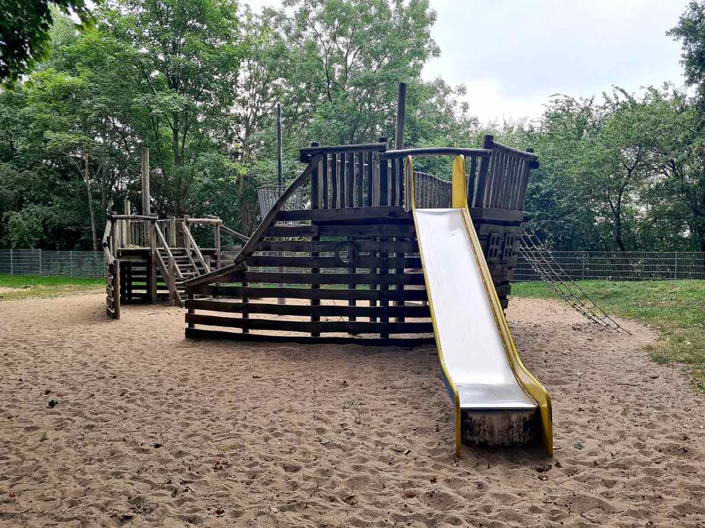 Großes hölzernes Piratenschiff-Spielgerät mit Rutsche, Seilnetz und Kletteraufgang, eingebettet in eine weitläufige Sandfläche und umgeben von Bäumen.
