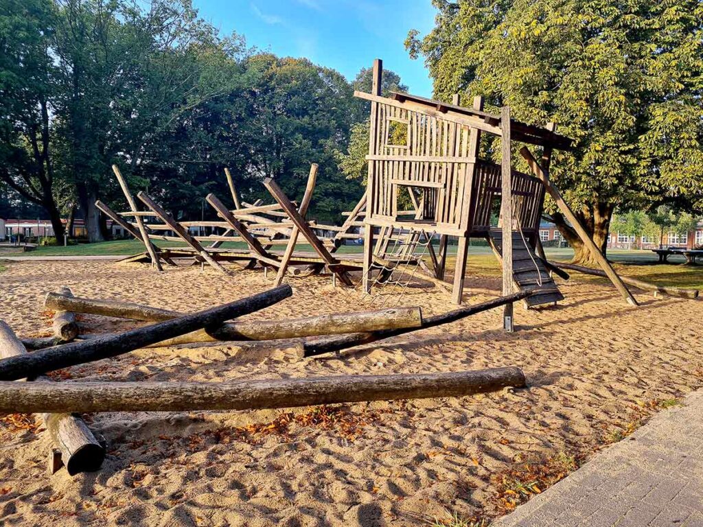Panoramaaufnahme des großen Abenteuerspielplatzes in Friedehorst. Im Vordergrund und Mittelgrund verlaufen zahlreiche schräge Holzbalken über den Sandboden, die als Balancier- oder Kletterelemente dienen. Rechts steht ein großes Holzspielhaus mit Rampe und Kletternetz. Im Hintergrund sind Wiesen, Bäume und Gebäude zu sehen.