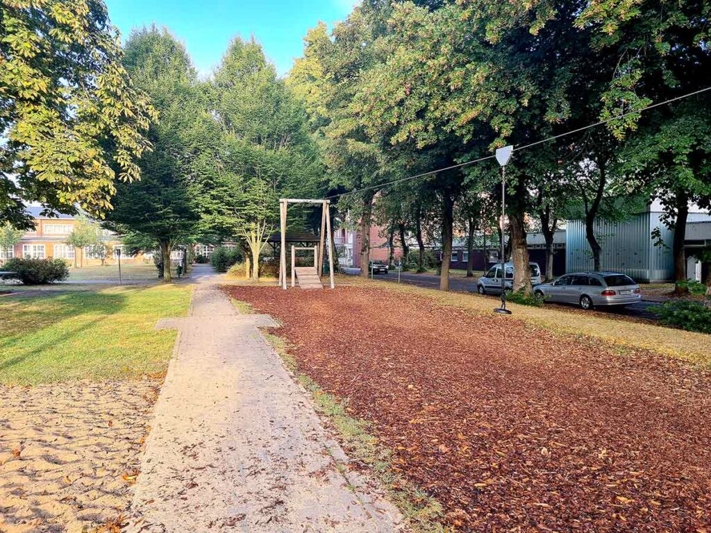 Blick auf die Seilrutsche des Spielplatzes Friedehorst. Rechts im Bild verläuft das Seil mit einem Tellergriff, darunter ein Bereich mit weichem Rindenmulch. Links führt ein gepflasterter Weg vorbei, gesäumt von Bäumen und Wiese. Im Hintergrund stehen Gebäude und parkende Autos.