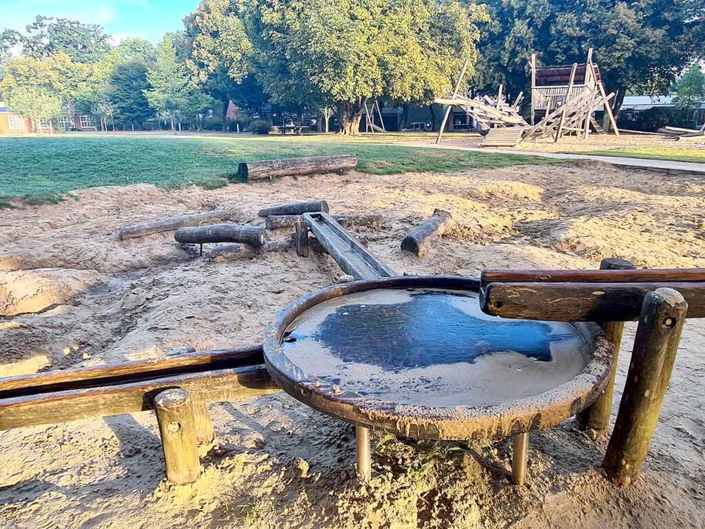 Wasserspielbereich auf dem Spielplatz Friedehorst. Im Vordergrund befindet sich ein rundes Wasserbecken aus Holz mit angeschlossenen Wasserläufen, die über den Sandboden verlaufen. Dahinter ist die große hölzerne Kletteranlage des Spielplatzes zu sehen.