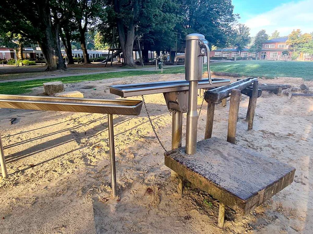 Handpumpe aus Edelstahl auf dem Spielplatz Friedehorst, befestigt auf einer erhöhten Holzplattform. Über eine Rinne aus Metall wird Wasser in einen Spielbereich mit Sand geleitet. Im Hintergrund sind grüne Wiese, Spielgeräte, große Bäume und Schulgebäude zu sehen.
