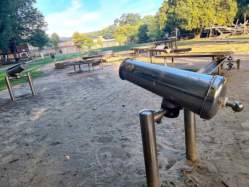 Edelstahl-Wasserkanonen auf dem Spielplatz Friedehorst, montiert auf stabilen Metallpfosten. Im Hintergrund sind weitere Spiel- und Wasserspielelemente sowie Bäume und Gebäude sichtbar.