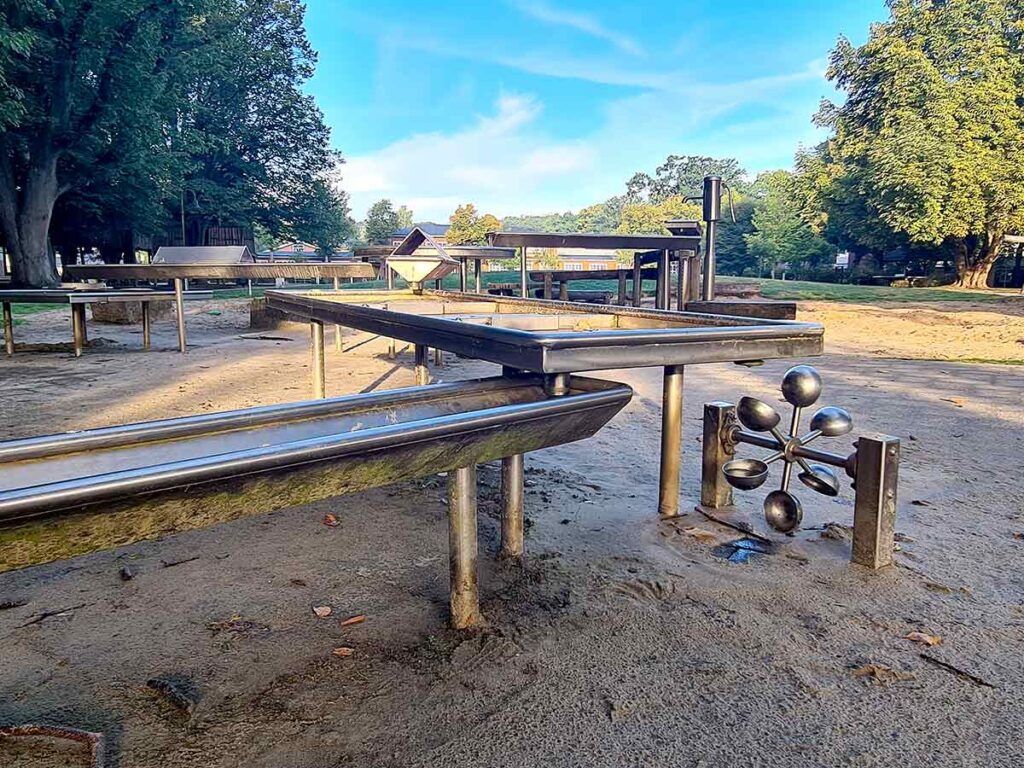 Edelstahl-Wasserspielanlage mit Rinnen, Becken und einem drehbaren Schaufelrad auf dem Spielplatz Friedehorst. Die Anlage steht auf festem Sandboden und ist von Bäumen sowie weiteren Spielgeräten umgeben. Ideal zum Experimentieren mit Wasser und Bewegung.