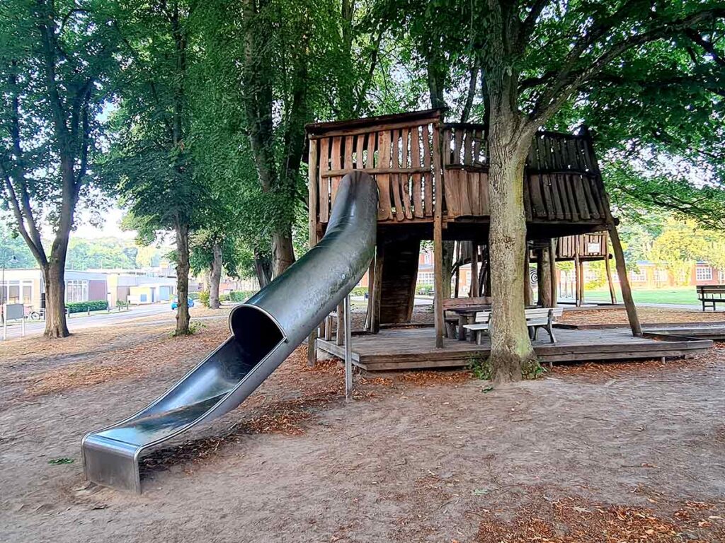 Große Holzspielstruktur mit überdachter Plattform und langer Tunnelrutsche auf dem Spielplatz Friedehorst. Die Anlage steht im Schatten hoher Bäume und bietet darunter Sitzmöglichkeiten mit Picknickbänken