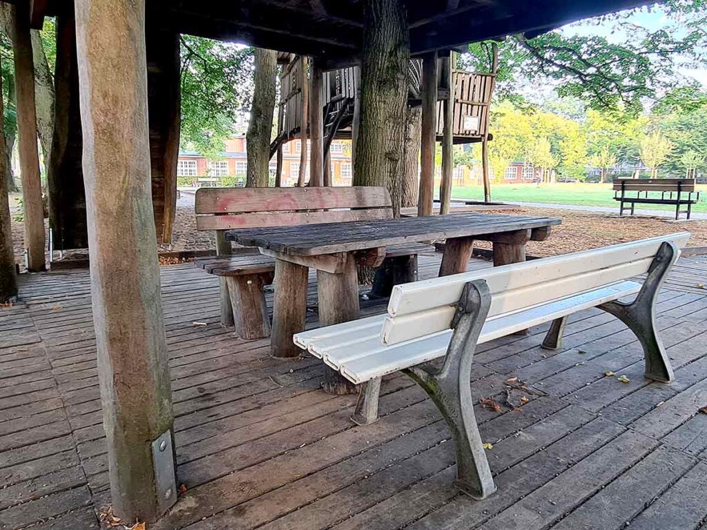 Sitzbereich mit rustikaler Holzsitzgruppe und weißer Parkbank unter einer großen Holzspielstruktur im Spielplatz Friedehorst.