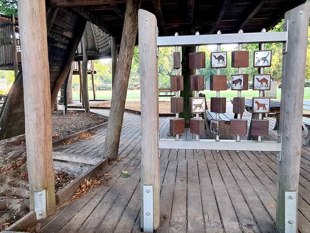 Drehspiel mit Holzwürfeln und Tiermotiven unter einer großen Holzspielstruktur. Die Würfel sind an Metallstangen befestigt und zeigen verschiedene Tierabbildungen wie Hund, Katze und Kamel. Die Konstruktion steht auf einem Holzdeck zwischen dicken Holzpfosten.