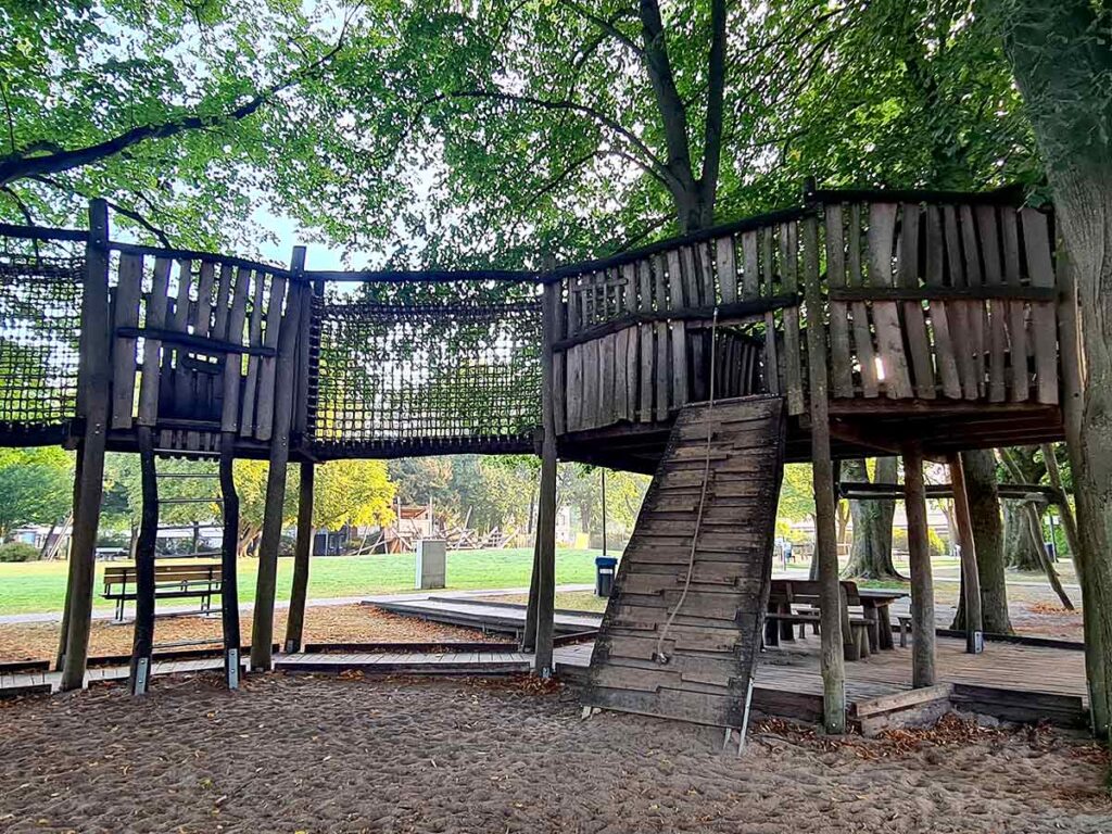 Große Holzspielstruktur mit schräger Kletterrampe, Kletternetz und erhöhter Plattform unter Bäumen. Darunter befindet sich ein Holzdeck mit Sitzgelegenheiten. Im Hintergrund sind Rasenflächen, Bänke und weitere Spielgeräte sichtbar.