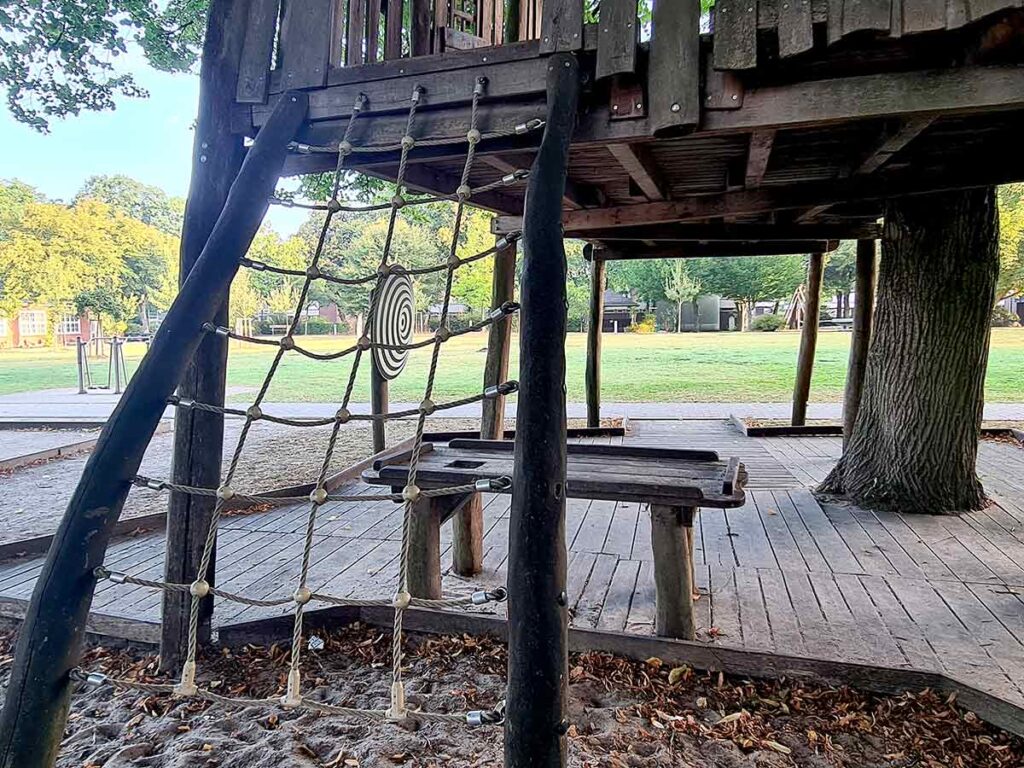 letternetz aus Seilen zwischen zwei schrägen Holzstämmen, befestigt an einer erhöhten Holzspielstruktur. Dahinter ein hölzerner Tisch mit Bank auf einem Holzdeck, daneben ein Baum. Im Hintergrund Rasenfläche und weitere Spielgeräte.