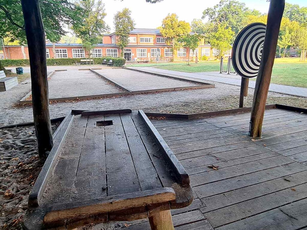 Holztisch mit eingelassener Spielfläche auf einem Holzdeck unter einer überdachten Spielstruktur. Rechts daneben eine große schwarz-weiß gestreifte Zielscheibe. Im Hintergrund mehrere rechteckige Sandflächen und ein Backsteingebäude mit weißen Fenstern.