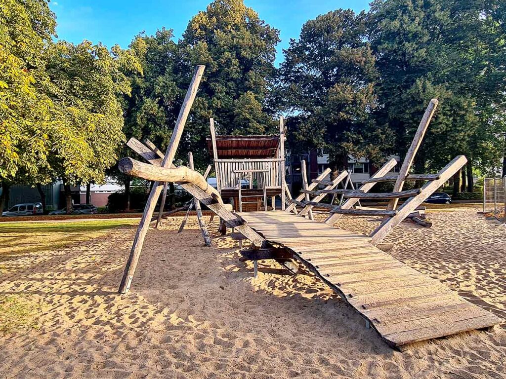 Großes Kletterspielgerät aus Holz auf dem Spielplatz in Friedehorst. Im Vordergrund eine breite Rampe, die zu einer erhöhten Plattform führt, umgeben von vielen schräg angeordneten Holzbalken und Seilen. Der gesamte Bereich ist mit Sand bedeckt.