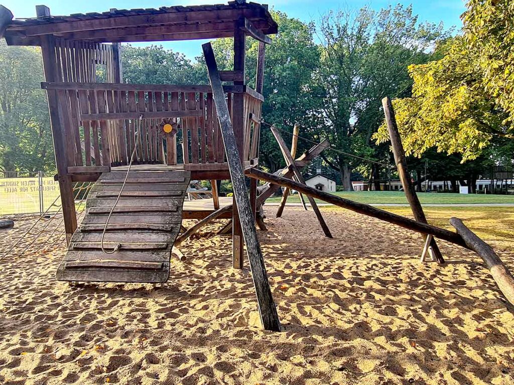 Seitliche Ansicht eines großen Holzspielgeräts auf dem Spielplatz Friedehorst. Eine schräge Rampe mit Kletterseil führt hinauf zur Plattform. Rundherum verlaufen schräge Kletterstangen und Seil-Balancierelemente. Der Boden ist vollständig mit Sand bedeckt, im Hintergrund sind Bäume und eine große Wiese zu sehen.