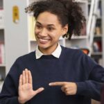 Eine junge, farbige Frau macht Handzeichen (Gebärdensprache) und lächelt. 