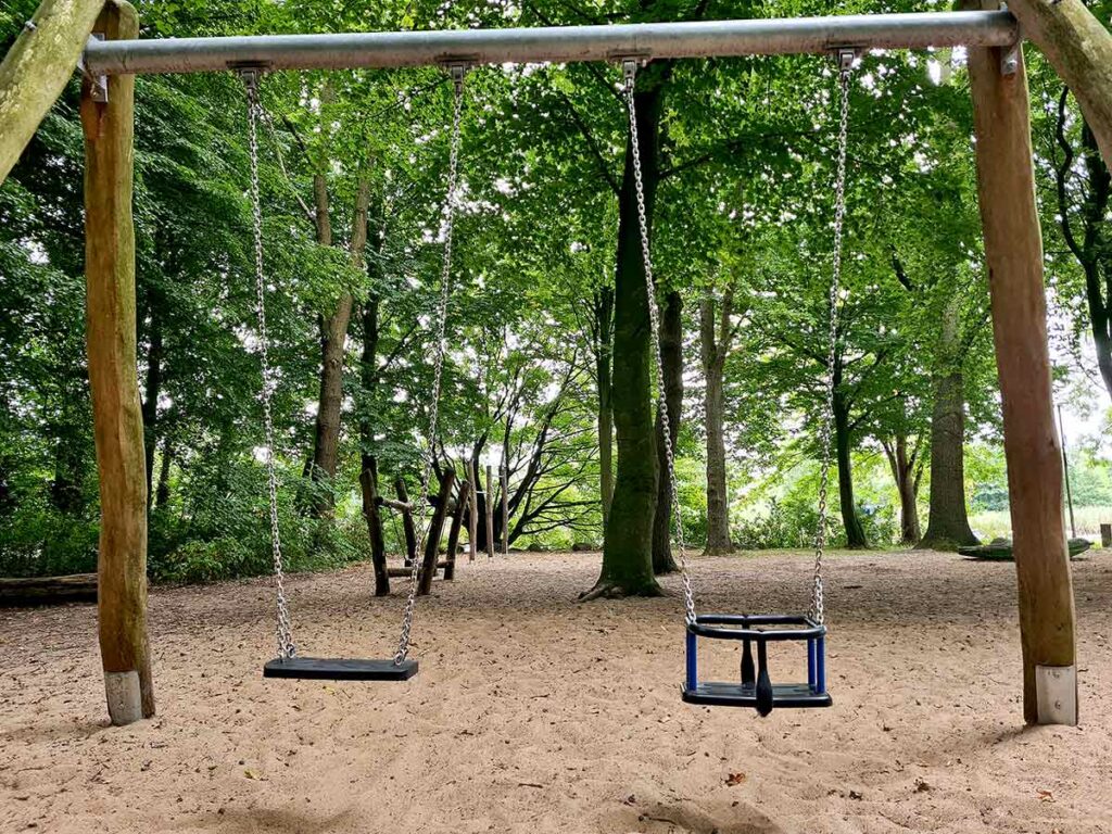 Zwei Schaukeln im Sandbereich eines Waldspielplatzes: Links eine klassische Sitzschaukel, rechts eine Kleinkindschaukel mit Sicherheitsbügel. Umgeben von hohen Bäumen und natürlicher Vegetation. Im Hintergrund sind weitere Holzspielgeräte zu erkennen.
